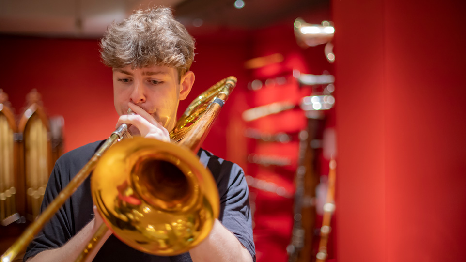 A male student playing the trumpet, with historical instruments on display in a red room, in the background.